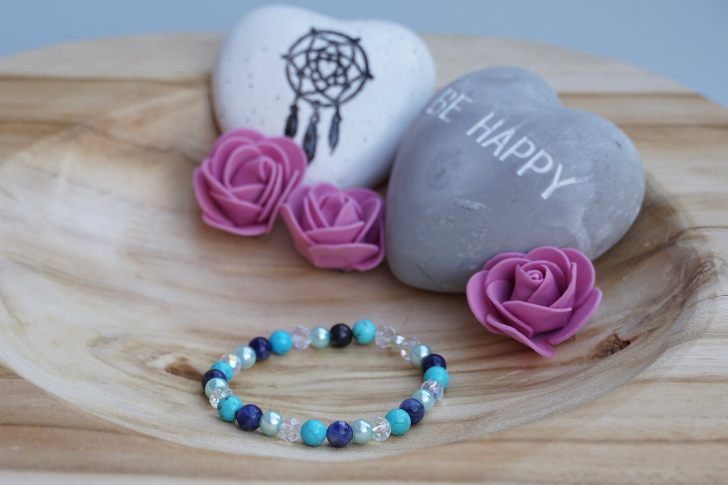 Rundes Armband aus Turquenit, Sodalith und hellblauer Acrylperlen und Glasperlen auf Holzschale, daneben zwei herzförmige Steine mit Schrift und Traumfänger sowie drei rosa Rosen aus Stoff.