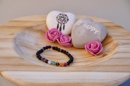 Armband aus schwarzen Lavaperlen mit sieben farbigen Chakrasteinen und kleinen Edelstahlzwischenringen auf Holzschale, daneben zwei herzförmige Steine mit Schrift und Traumfänger sowie drei rosa Rosen aus Stoff.