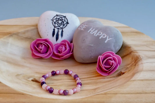 Rundes Armband aus Rosenquarz, Amethyst und rosa Acrylperlen auf Holzschale, daneben zwei herzförmige Steine mit Schrift und Traumfänger sowie drei rosa Rosen aus Stoff.