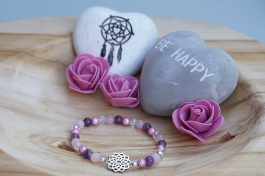 Rundes Armband aus Rosenquarz, Amethyst und rosa Acrylperlen mit Glasperlen und Blume des Lebens-Anhänger auf Holzschale, daneben zwei herzförmige Steine mit Schrift und Traumfänger sowie drei rosa Rosen aus Stoff.