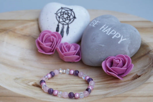 Rundes Armband aus Rosenquarz, Amethyst und rosa Acrylperlen mit Glasperlen auf Holzschale, daneben zwei herzförmige Steine mit Schrift und Traumfänger sowie drei rosa Rosen aus Stoff.
