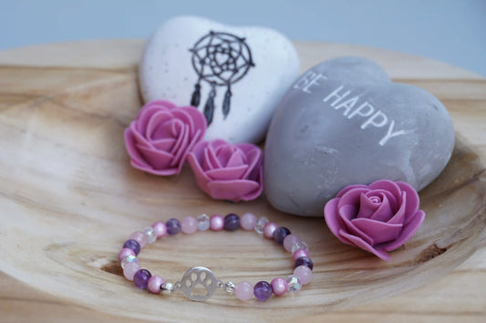 Rundes Armband aus Rosenquarz, Amethyst und rosa Acrylperlen mit Glasperlen und Hundepfoten-Anhänger auf Holzschale, daneben zwei herzförmige Steine mit Schrift und Traumfänger sowie drei rosa Rosen aus Stoff.
