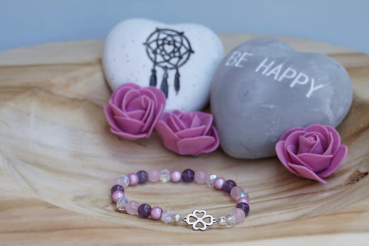 Rundes Armband aus Rosenquarz, Amethyst und rosa Acrylperlen mit Glasperlen und Kleeblatt-Anhänger auf Holzschale, daneben zwei herzförmige Steine mit Schrift und Traumfänger sowie drei rosa Rosen aus Stoff.