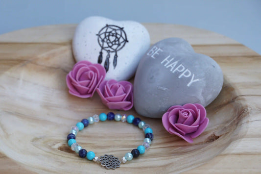 Rundes Armband aus Turquenit, Sodalith und hellblauer Acrylperlen sowie Glasperlen mit Blume des Lebens-Anhänger auf Holzschale, daneben zwei herzförmige Steine mit Schrift und Traumfänger sowie drei rosa Rosen aus Stoff.
