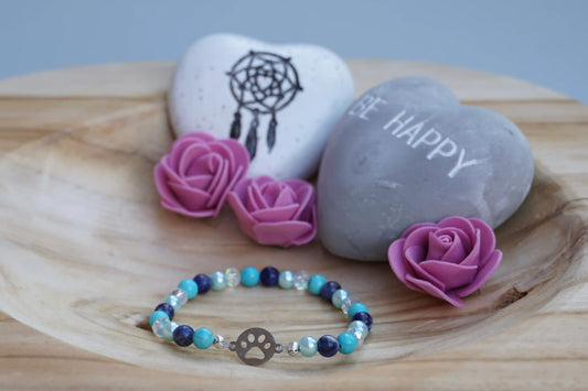 Rundes Armband aus Turquenit, Sodalith und hellblauer Acrylperlen sowie Glasperlen mit Hundepfoten-Anhänger auf Holzschale, daneben zwei herzförmige Steine mit Schrift und Traumfänger sowie drei rosa Rosen aus Stoff.