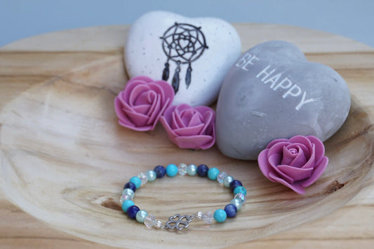 Rundes Armband aus Turquenit, Sodalith und hellblauer Acrylperlen sowie Glasperlen mit Kleeblatt-Anhänger auf Holzschale, daneben zwei herzförmige Steine mit Schrift und Traumfänger sowie drei rosa Rosen aus Stoff.