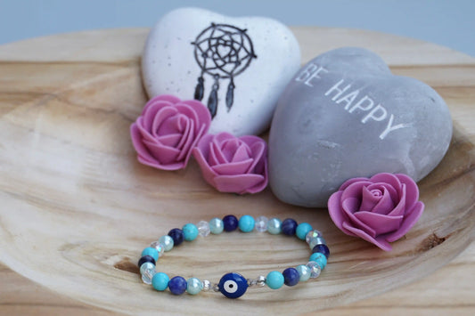 Rundes Armband aus Turquenit, Sodalith und hellblauer Acrylperlen sowie Glasperlen mit Nazaramulett-Anhänger auf Holzschale, daneben zwei herzförmige Steine mit Schrift und Traumfänger sowie drei rosa Rosen aus Stoff.