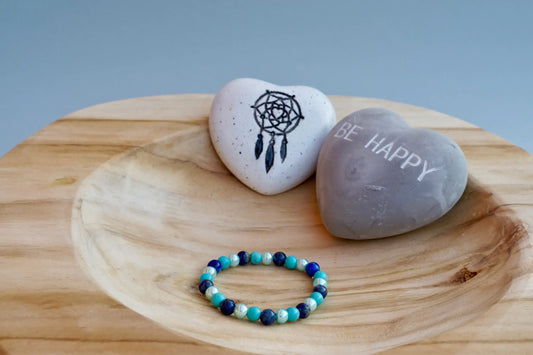 Rundes Armband aus Turquenit, Sodalith und hellblauer Acrylperlen auf Holzschale, daneben zwei herzförmige Steine mit Schrift und Traumfänger sowie drei rosa Rosen aus Stoff.