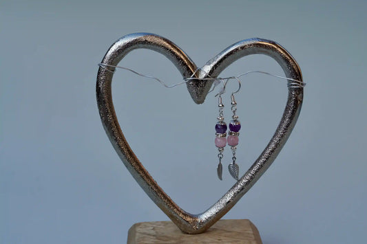Ohrringe mit silberfarbenen Haken, zwei Edelsteinperlen in Amethyst und Rosenquarz sowie einem silberfarbenen Blattanhänger aufgehängt an einem herzförmigen Metallständer.