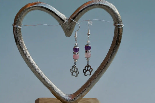 Ohrringe mit silberfarbenen Haken, zwei Edelsteinperlen in Amethyst und Rosenquarz sowie einem silberfarbenen Hundepfotenanhänger aufgehängt an einem herzförmigen Metallständer.