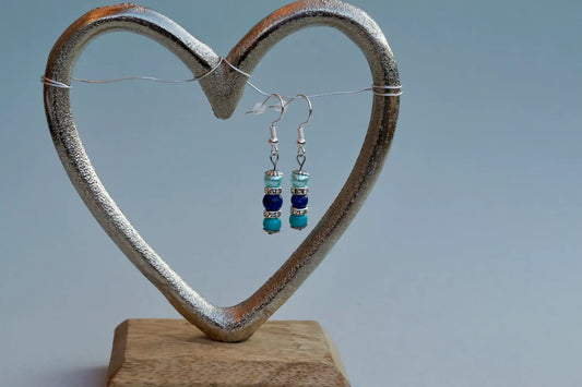 Ohrringe mit silberfarbenen Haken, zwei Edelsteinperlen in Turquenit und Sodalith sowie hellblauer Acrylperle aufgehängt an einem herzförmigen Metallständer.