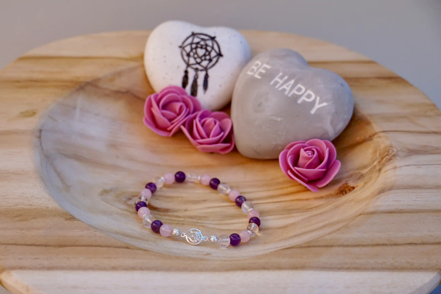 Edelsteinsarmband mit Rosenquarz, Amethyst und Bergkristall auf Holzschale, daneben zwei herzförmige Steine mit Schrift und Traumfänger sowie drei rosa Rosen aus Stoff.