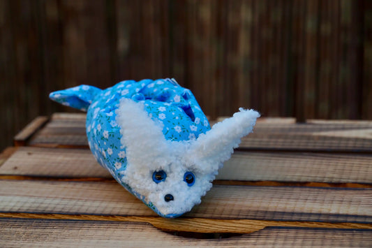 Kuscheltier in Form eines weißen Hasenkopfes mit Knopfaugen, eingewickelt in blauem Blümchen Stoff auf Holztisch