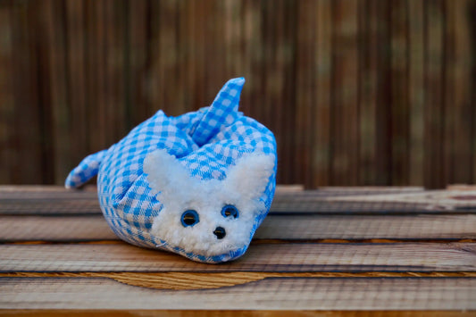 Kuscheltier in Form eines weißen Hasenkopfes mit blauen Knopfaugen, eingewickelt in blau-weiß karierten Stoff, auf Holztisch