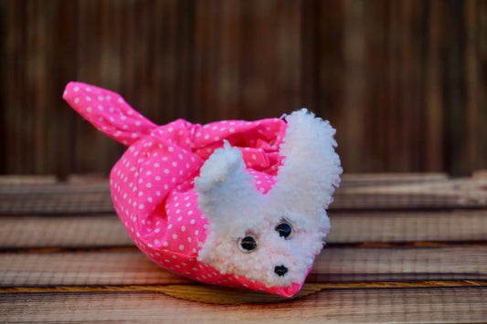 Kuscheltier in Form eines weißen Hasenkopfes mit schwarzen Knopfaugen, eingewickelt in pinkem Stoff mit weißen Punkten, auf Holztisch