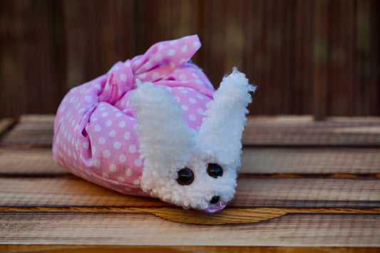 Kuscheltier in Form eines weißen Hasenkopfes mit schwarzen Knopfaugen, eingewickelt in rosa Stoff mit weißen Punkten, auf Holztisch