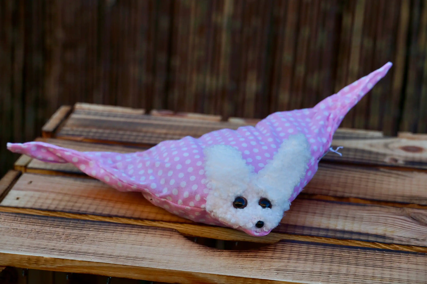 Kuscheltier in Form eines weißen Hasenkopfes mit schwarzen Knopfaugen, eingewickelt in rosa Stoff mit weißen Punkten, auf Holztisch