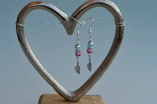 Ohrringe mit silberfarbenen Haken, zwei Perlen in Türkis und Rosa sowie einem silberfarbenen Blattanhänger aufgehängt an einem herzförmigen Metallständer.