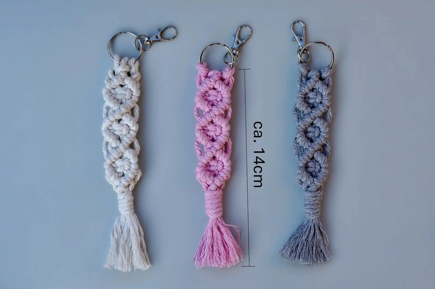 Drei Makramee-Schlüsselanhänger in Weiß, Rosa und Grau mit Knotenmustern und Fransen, jeweils mit Metallring und Karabinerhaken