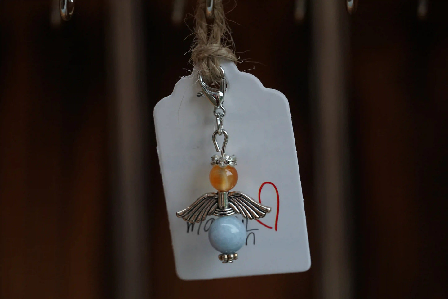 Anhänger in Form eines Schutzengels mit Karneol- und Chalcedonperlen, silberfarbene Flügel, an einem Band befestigt.