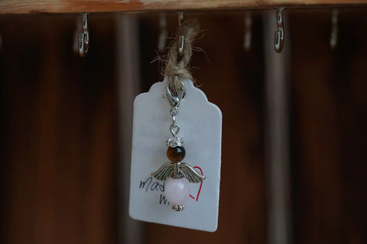 Anhänger in Form eines Schutzengels mit Tigerauge- und Rosenquarzperlen, silberfarbene Flügel, an einem Band befestigt.