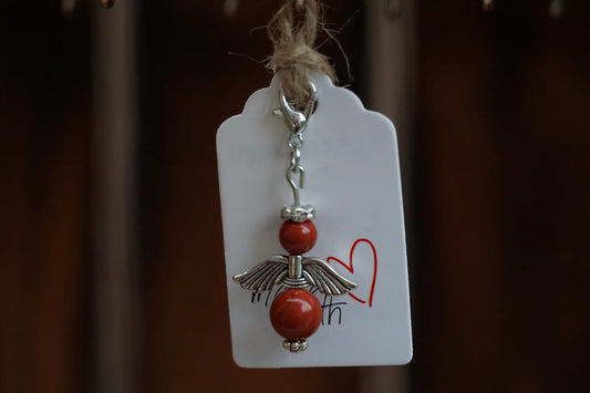 Anhänger in Form eines Schutzengels mit roten Jaspisperlen, silberfarbene Flügel, an einem Band befestigt.