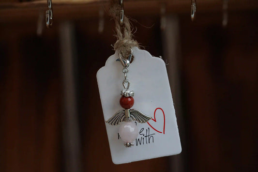 Anhänger in Form eines Schutzengels mit roter Jaspis- und Rosenquarzperlen, silberfarbene Flügel, an einem Band befestigt.