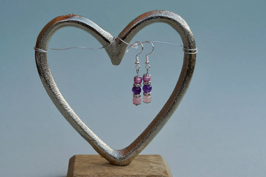 Ohrringe mit silberfarbenen Haken, zwei Edelsteinperlen in Amethyst und Rosenquarz, sowie rosa Acrylperle aufgehängt an einem herzförmigen Metallständer.