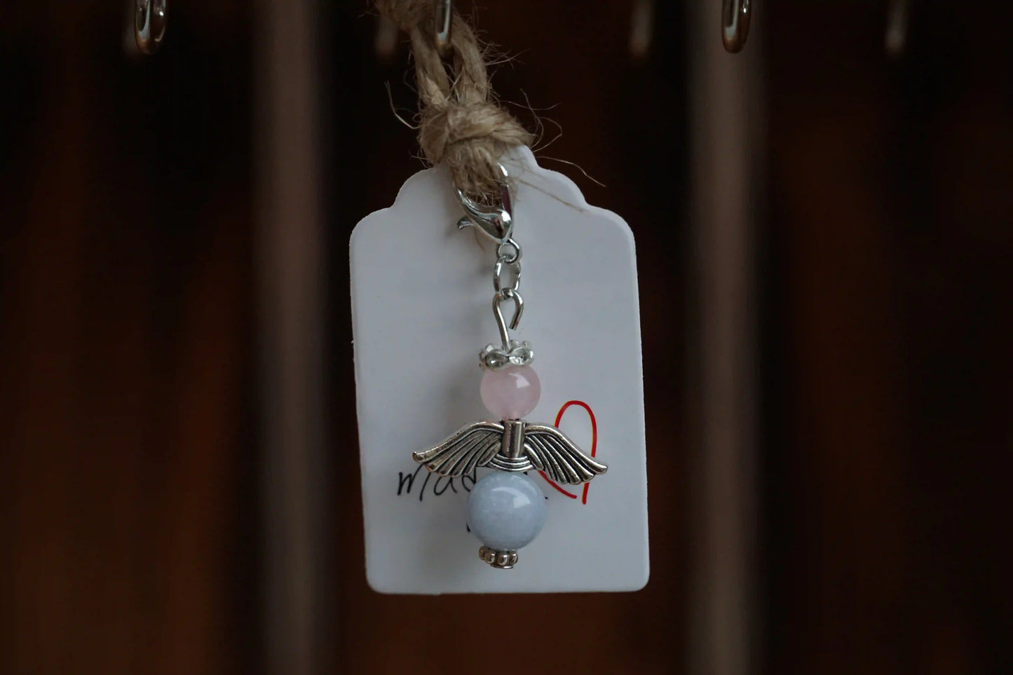 Anhänger in Form eines Schutzengels mit Rosenquarz- und Chalcedonperlen, silberfarbene Flügel, an einem Band befestigt.
