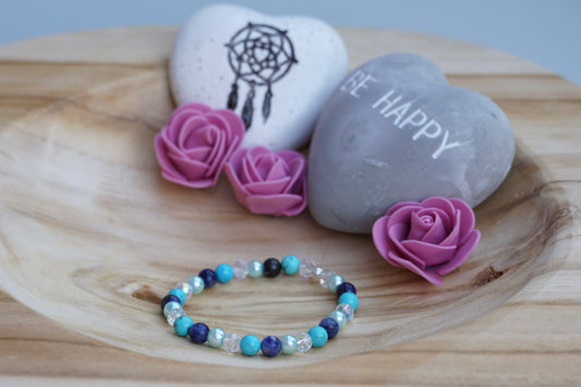 Rundes Armband aus Turquenit, Sodalith und hellblauer Acrylperlen und Glasperlen auf Holzschale, daneben zwei herzförmige Steine mit Schrift und Traumfänger sowie drei rosa Rosen aus Stoff.