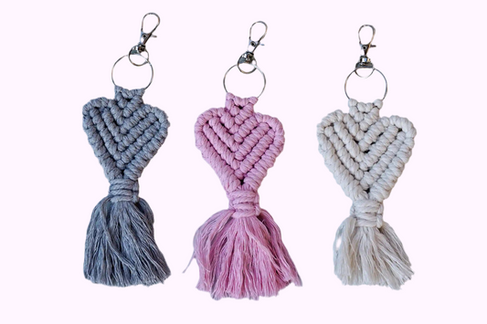 Drei Makramee-Schlüsselanhänger in Grau, Rosa und weiß mit Herzknotenmuster und Fransen, jeweils mit Metallring und Karabinerhaken