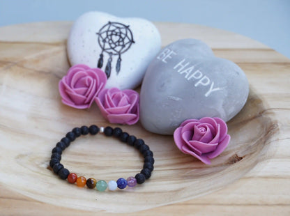 Armband aus schwarzen Lavaperlen mit sieben farbigen Chakrasteinen auf Holzschale, daneben zwei herzförmige Steine mit Schrift und Traumfänger sowie drei rosa Rosen aus Stoff.