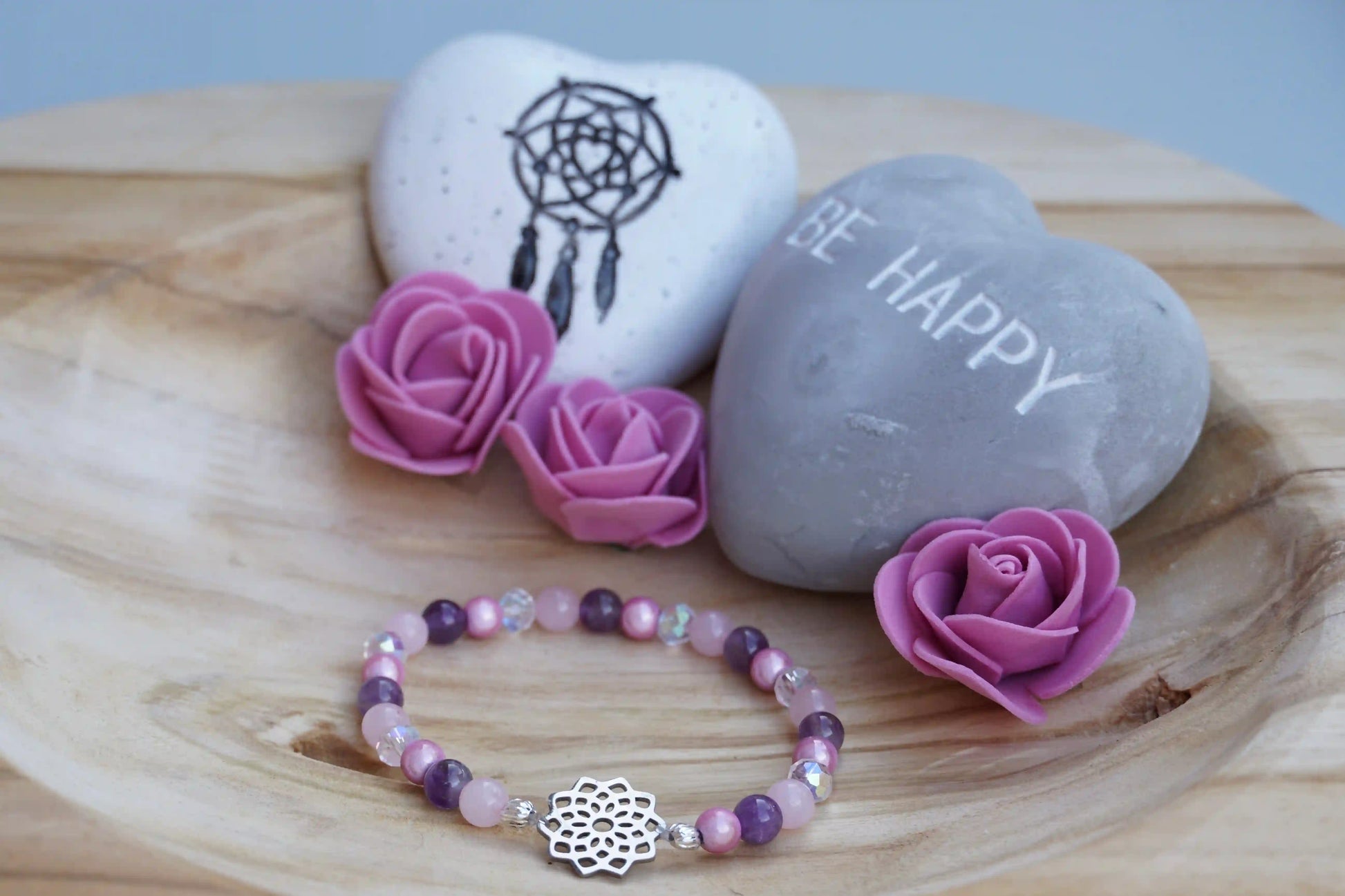 Rundes Armband aus Rosenquarz, Amethyst und rosa Acrylperlen mit Glasperlen und Blume des Lebens-Anhänger auf Holzschale, daneben zwei herzförmige Steine mit Schrift und Traumfänger sowie drei rosa Rosen aus Stoff.
