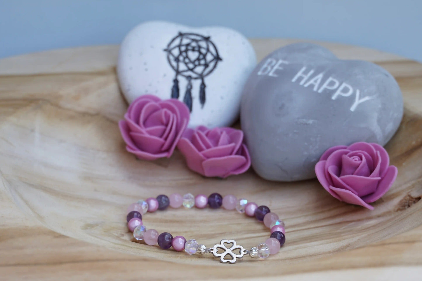 Rundes Armband aus Rosenquarz, Amethyst und rosa Acrylperlen mit Glasperlen und Kleeblatt-Anhänger auf Holzschale, daneben zwei herzförmige Steine mit Schrift und Traumfänger sowie drei rosa Rosen aus Stoff.