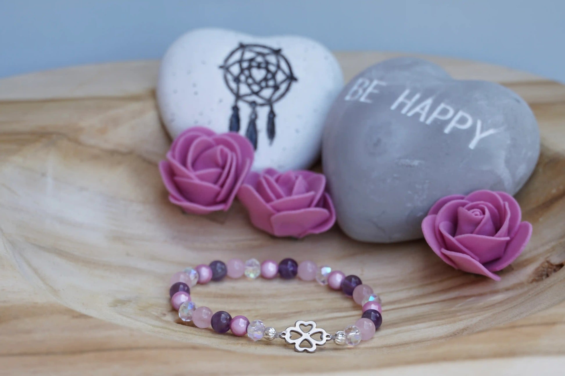 Rundes Armband aus Rosenquarz, Amethyst und rosa Acrylperlen mit Glasperlen und Kleeblatt-Anhänger auf Holzschale, daneben zwei herzförmige Steine mit Schrift und Traumfänger sowie drei rosa Rosen aus Stoff.