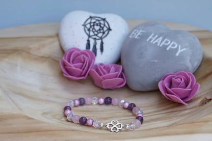 Rundes Armband aus Rosenquarz, Amethyst und rosa Acrylperlen mit Glasperlen und Kleeblatt-Anhänger auf Holzschale, daneben zwei herzförmige Steine mit Schrift und Traumfänger sowie drei rosa Rosen aus Stoff.