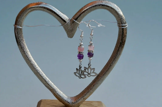 Ohrringe mit silberfarbenen Haken, zwei Edelsteinperlen in Amethyst und Rosenquarz sowie einem silberfarbenen Lotusblumenanhänger aufgehängt an einem herzförmigen Metallständer.