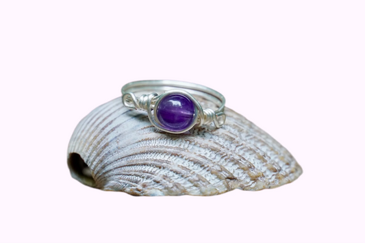 Ring mit einer Amethyst Edelsteinperle eingefasst in versilbertem Kupferdraht, platziert auf einer Muschel