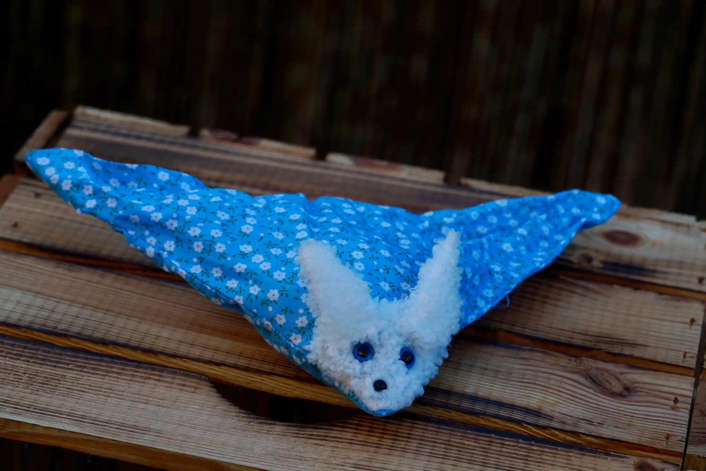 Kuscheltier in Form eines weißen Hasenkopfes mit Knopfaugen, eingewickelt in blauem Blümchen Stoff auf Holztisch
