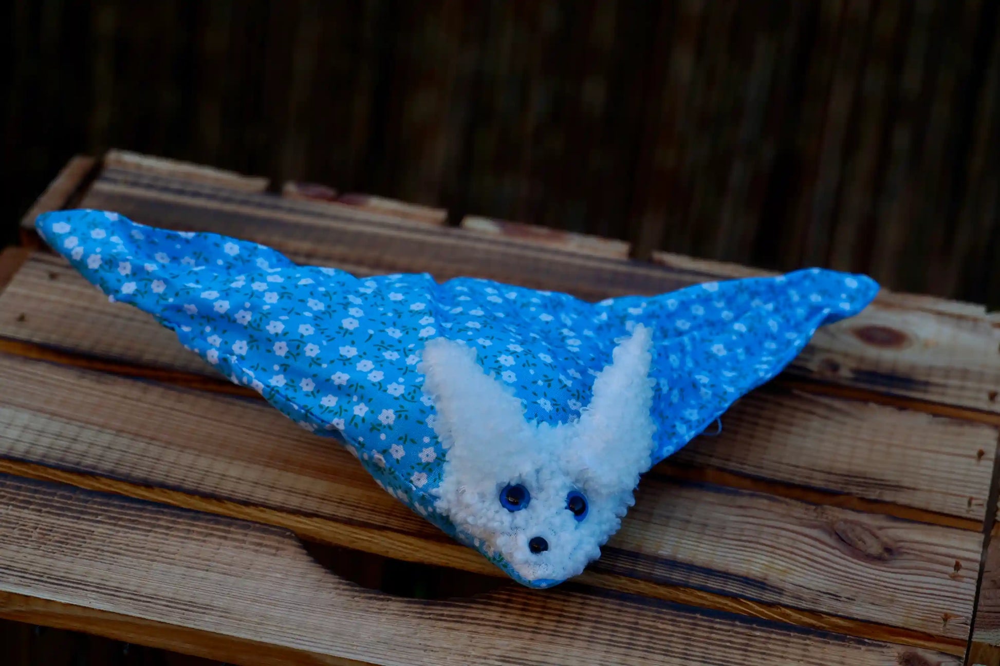 Kuscheltier in Form eines weißen Hasenkopfes mit Knopfaugen, eingewickelt in blauem Blümchen Stoff auf Holztisch