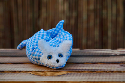 Kuscheltier in Form eines weißen Hasenkopfes mit blauen Knopfaugen, eingewickelt in blau-weiß karierten Stoff, auf Holztisch
