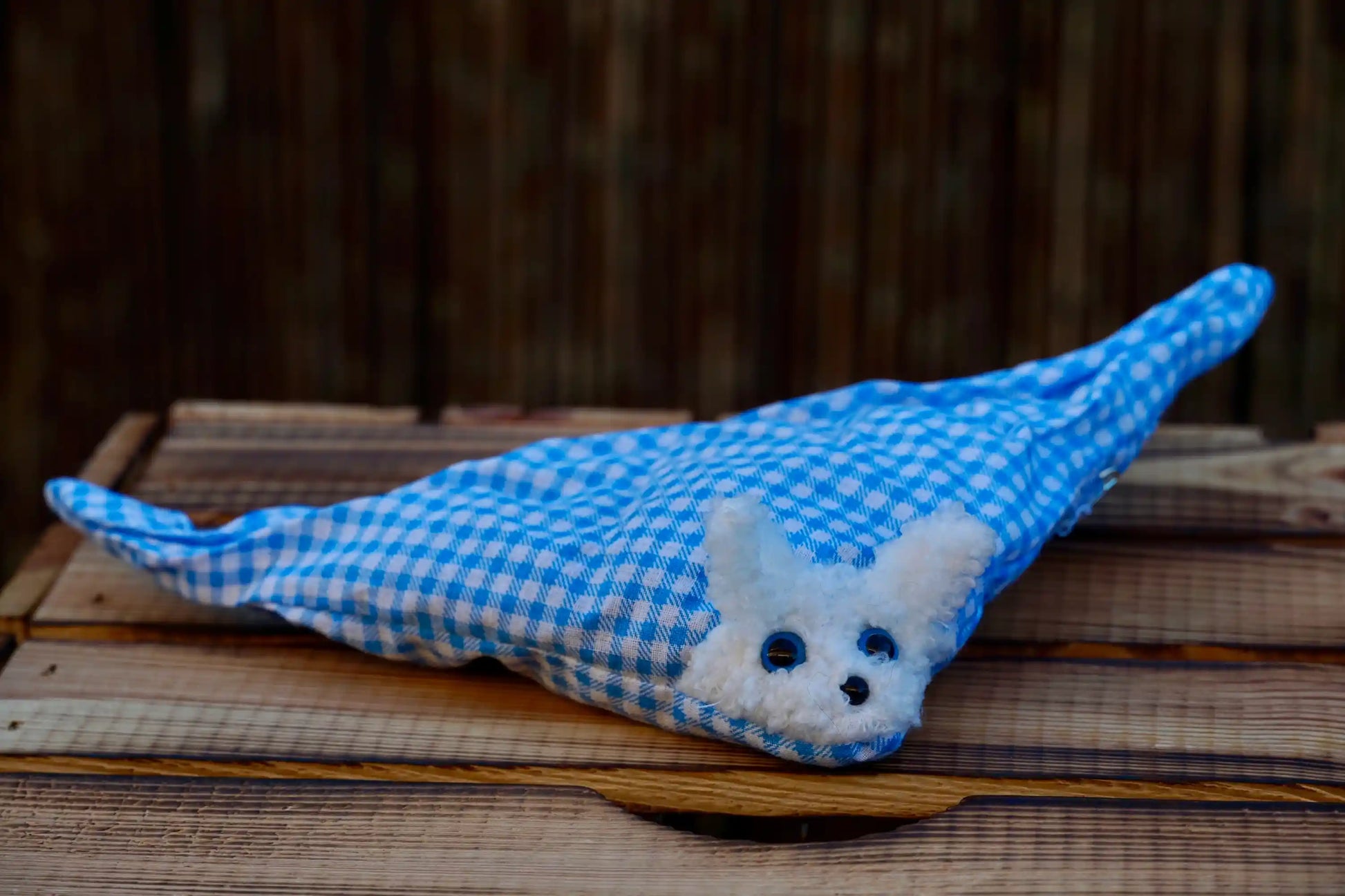 Kuscheltier in Form eines weißen Hasenkopfes mit blauen Knopfaugen, eingewickelt in blau-weiß karierten Stoff, auf Holztisch