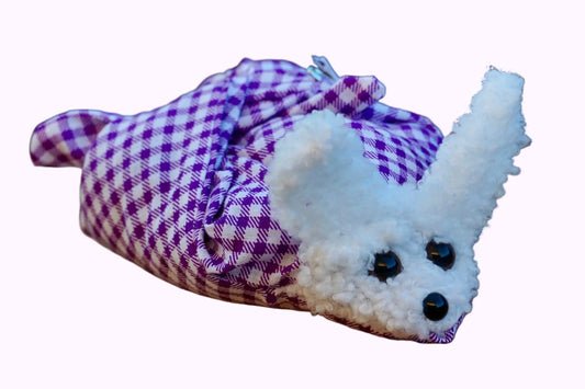 Kuscheltier in Form eines weißen Hasenkopfes mit Knopfaugen, eingewickelt in lila weiß kariertem Stoff, auf Holztisch