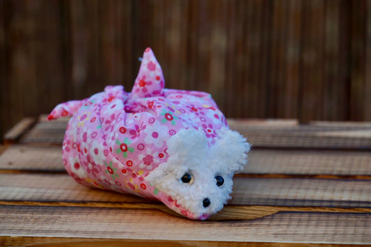 Kuscheltier in Form eines weißen Hasenkopfes mit Knopfaugen, eingewickelt in rosa  Blümchenstoff, auf Holztisch