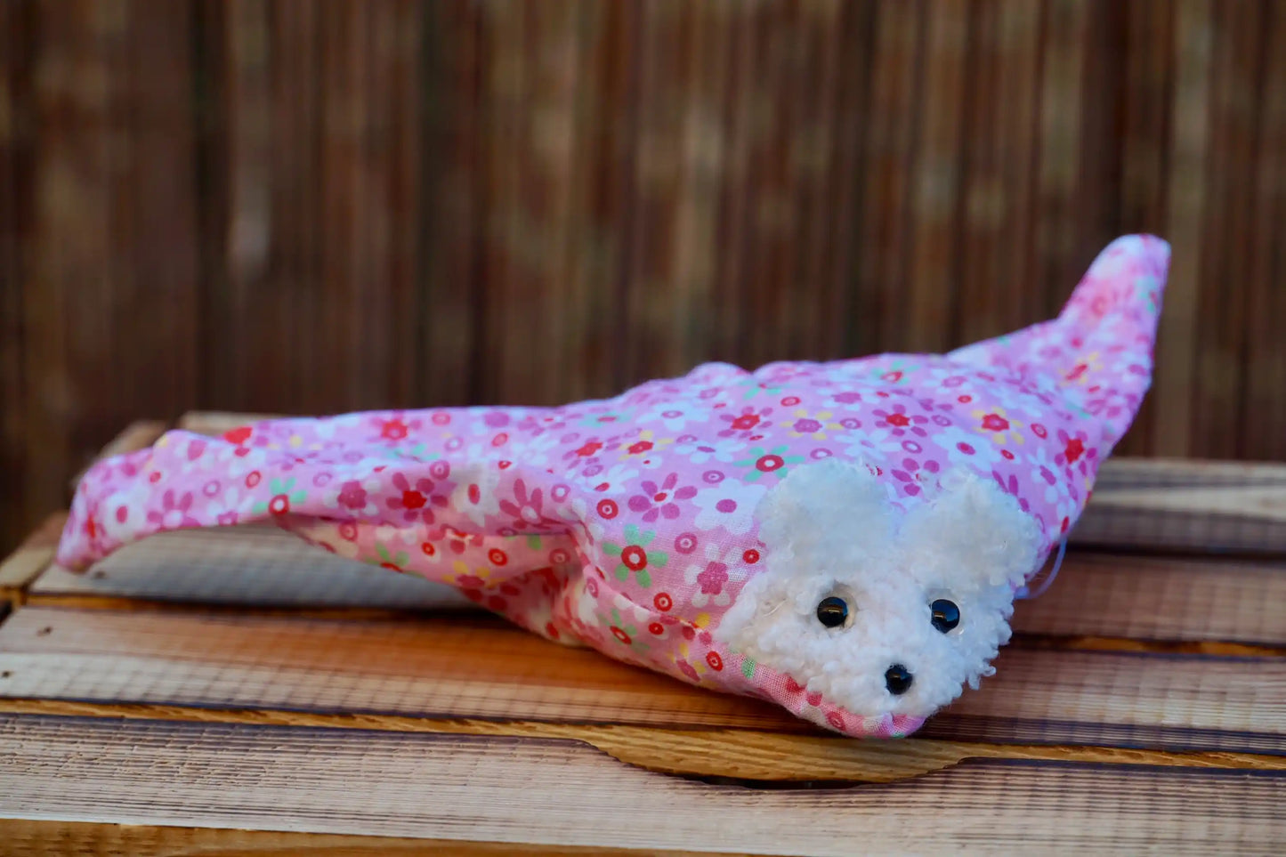 Kuscheltier in Form eines weißen Hasenkopfes mit Knopfaugen, eingewickelt in rosa  Blümchenstoff, auf Holztisch