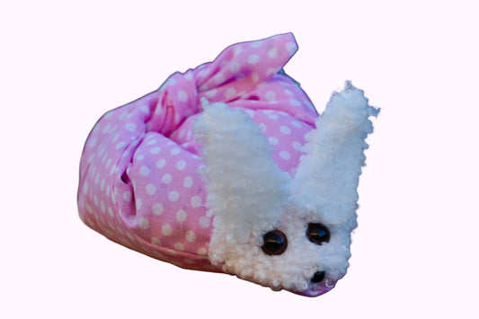 Kuscheltier in Form eines weißen Hasenkopfes mit schwarzen Knopfaugen, eingewickelt in rosa Stoff mit weißen Punkten
