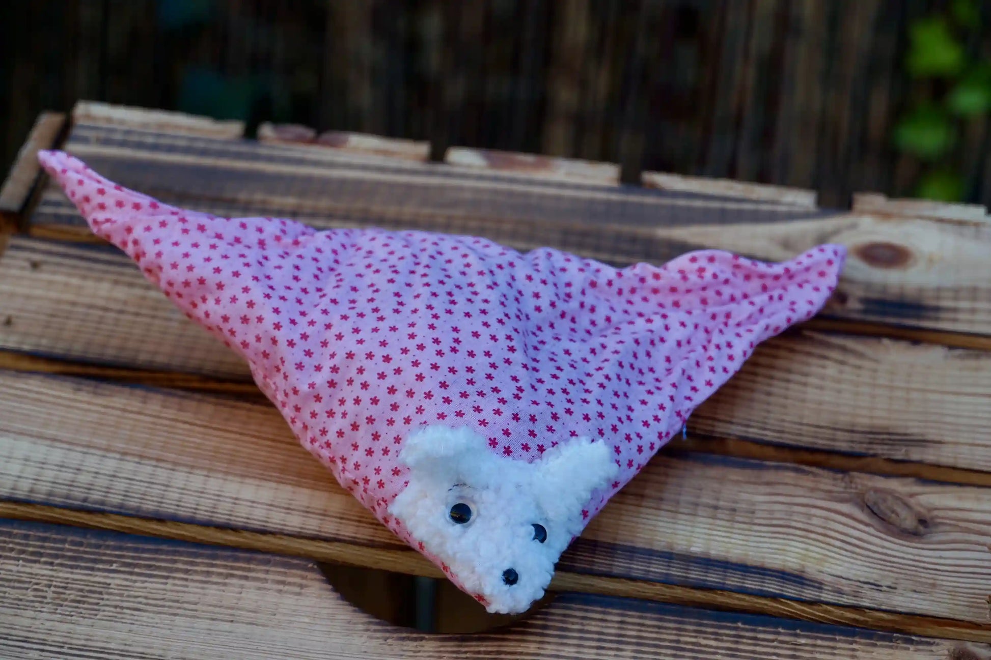 Kuscheltier in Form eines weißen Hasenkopfes mit Knopfaugen, eingewickelt in rosa Stoff mit Sternen auf Holztisch