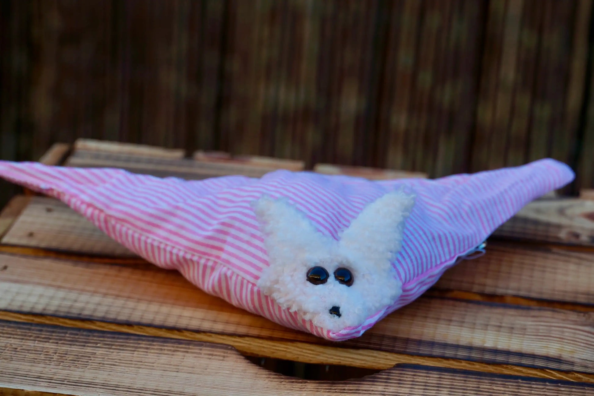 Kuscheltier in Form eines weißen Hasenkopfes mit Knopfaugen, eingewickelt in rosa weiß gestreiften Stoff, auf Holztisch