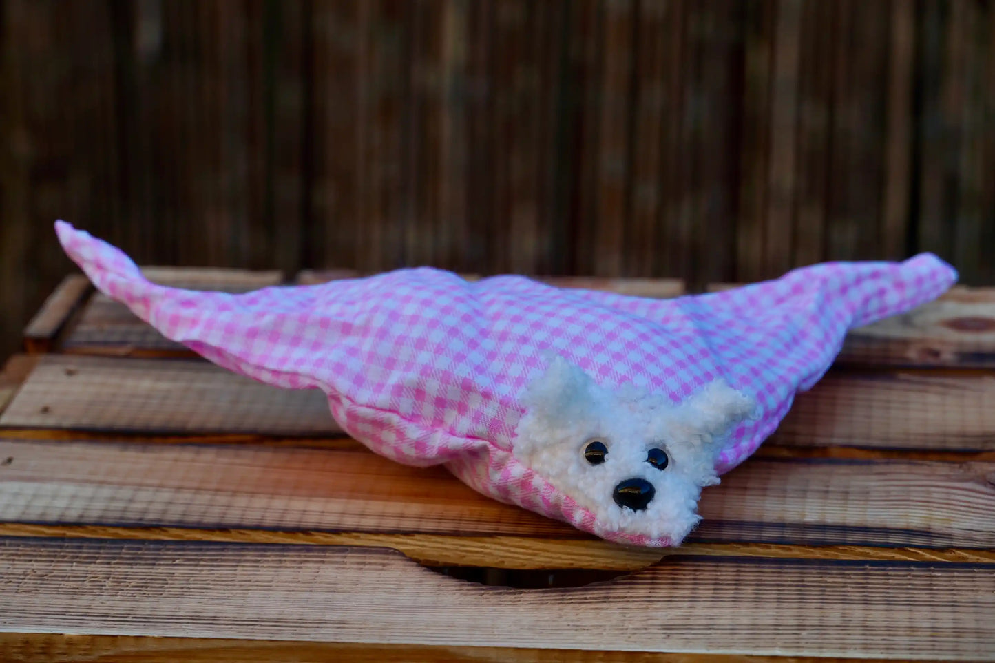 Kuscheltier in Form eines weißen Hasenkopfes mit Knopfaugen, eingewickelt in rosa-weiß karierten Stoff, auf Holztisch