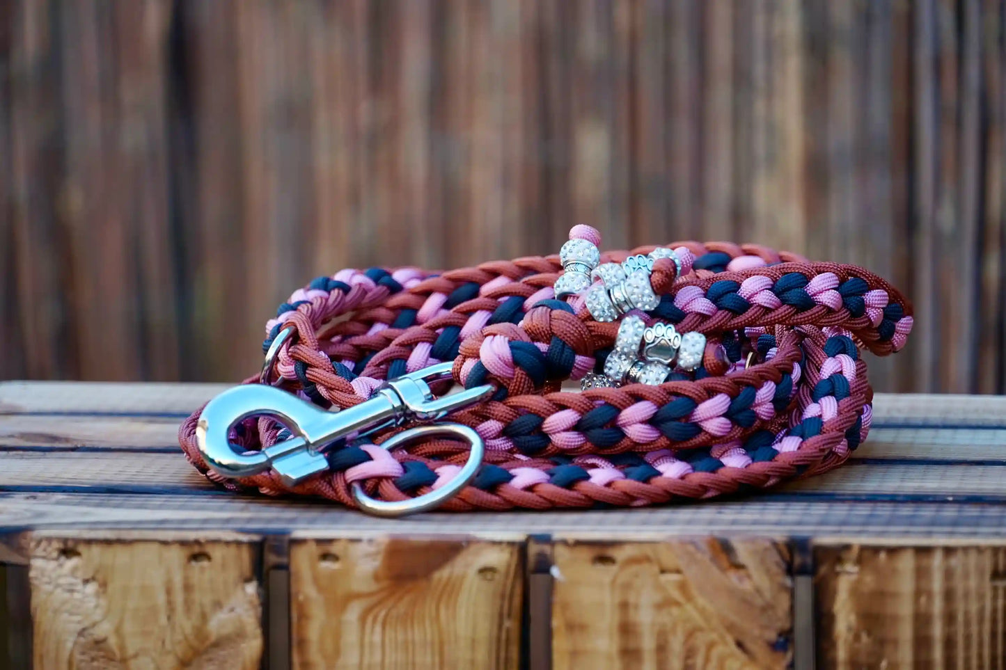 Geflochtene Paracord Hundeleine in Bronze, rosa und Anthrazittönen mit Metallkarabiner und Schmuckperlen am Griff auf holzkiste